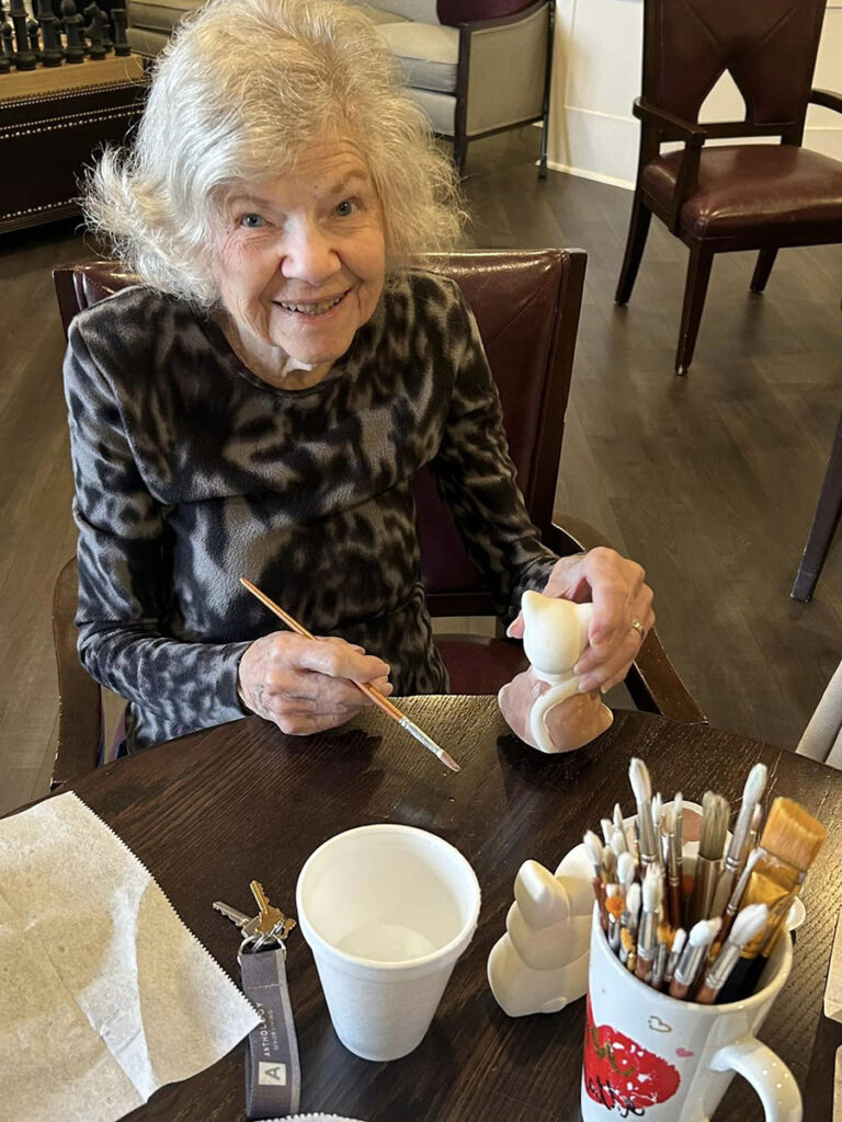A cheerful senior woman enjoys a craft event in a senior living community, amidst painted ceramic figures.