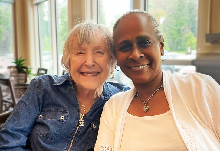 Two women smiling and sitting closely together in a bright room, symbolizing friendship, connection, and warmth.