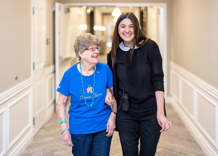 A smiling senior resident in a blue shirt walks down the hallway arm-in-arm with a younger female staff member, both enjoying a lighthearted moment at the senior living community.