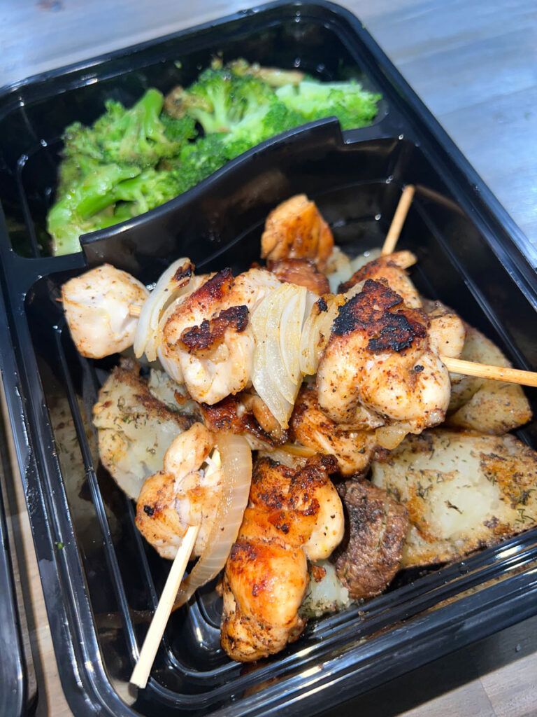 Grilled chicken skewers with roasted onions and potatoes, served alongside steamed broccoli in a black meal prep container on a wooden table.