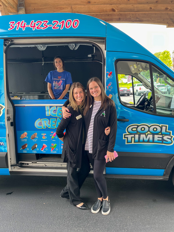 Two smiling women stand in front of an ice cream truck, one with her arm around the other. An employee inside the truck stands ready to serve.