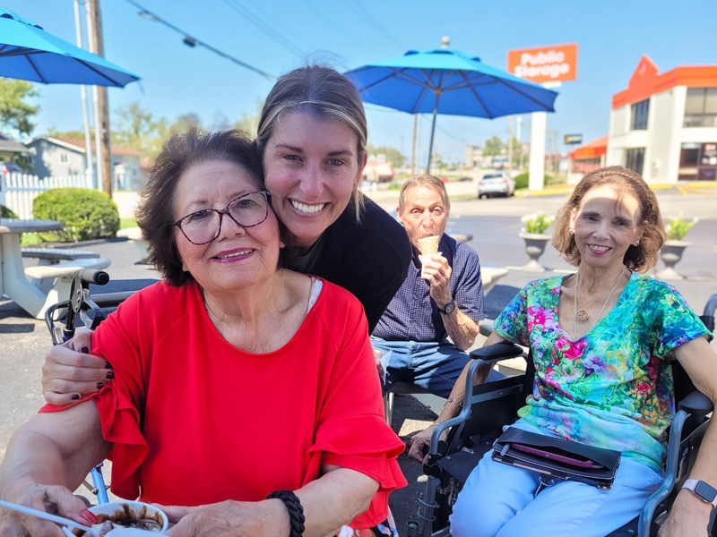 A group of senior residents enjoying ice cream outdoors under blue umbrellas, accompanied by a smiling staff member hugging one of the residents.