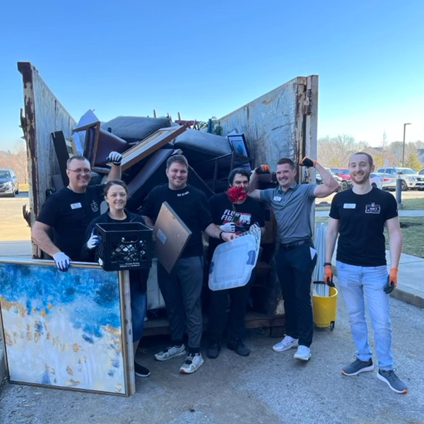 A diverse group of employees people standing in front of a large dumpster, smiling and holding up items from cleaning events.
