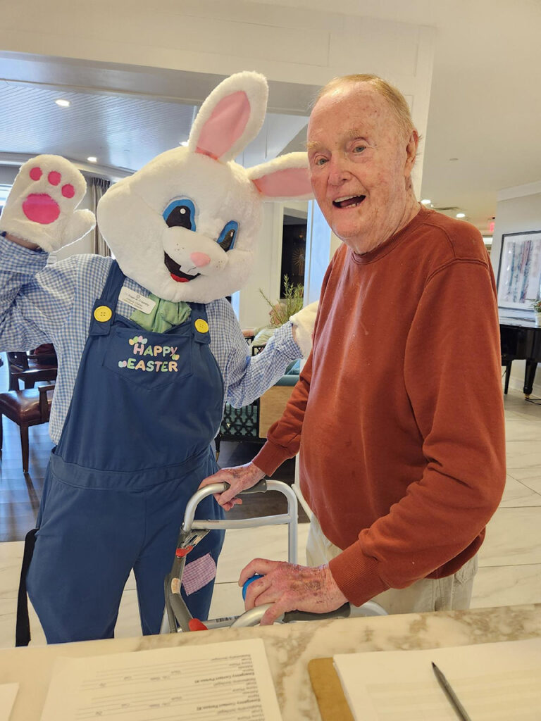 An elderly man in a burnt orange sweatshirt smiles with Hoppy the Easter Bunny at the front desk.