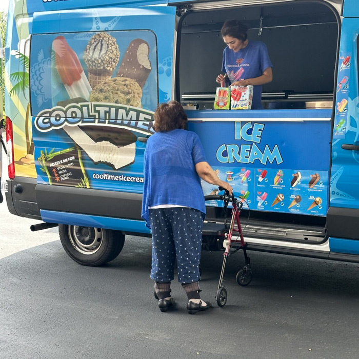 A senior resident uses a walker to approach an ice cream truck, choosing a treat on a sunny day, creating a joyful and nostalgic moment.