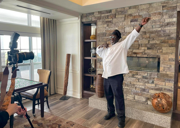 Chef playfully strikes a pose while being photographed in front of a stone fireplace, showcasing his vibrant personality and passion within the senior living community setting.