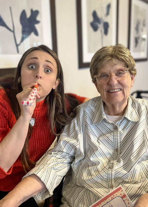 Senior resident smiles while seated next to a playful team member in a red sweater blowing a party horn, creating a fun and cheerful atmosphere during a community celebration.