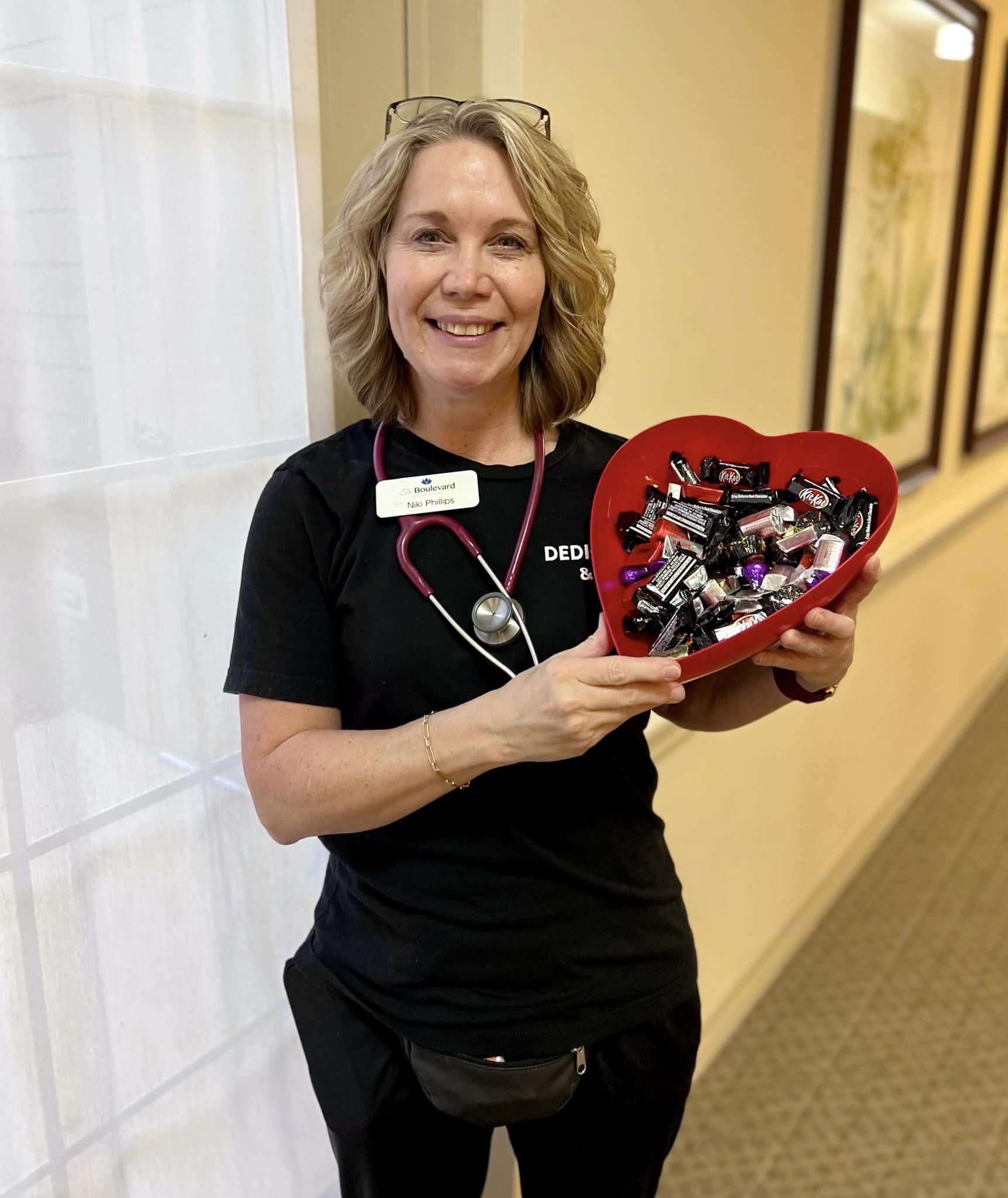 Smiling nurse holding a heart-shaped bowl filled with candy, offering treats to brighten the day of senior residents at a senior living community, promoting care and kindness.