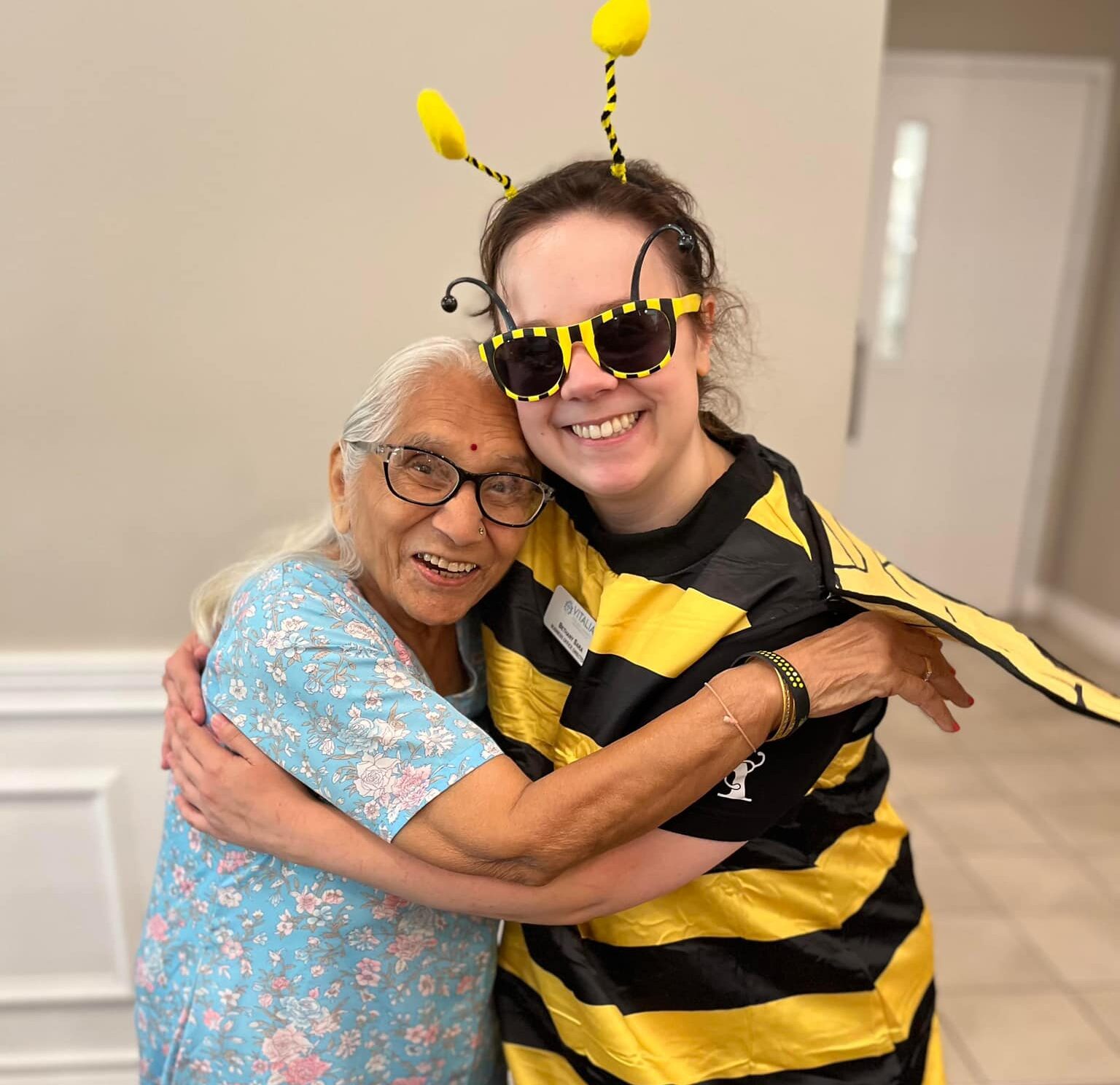 Senior resident in a floral dress hugs a smiling team member dressed in a bee costume with yellow antennae and glasses, creating a heartwarming moment at a community event.