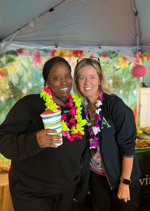 Two senior living team members enjoy a tropical-themed event, wearing colorful leis and holding a snow cone, standing in front of a festive backdrop at a community celebration.