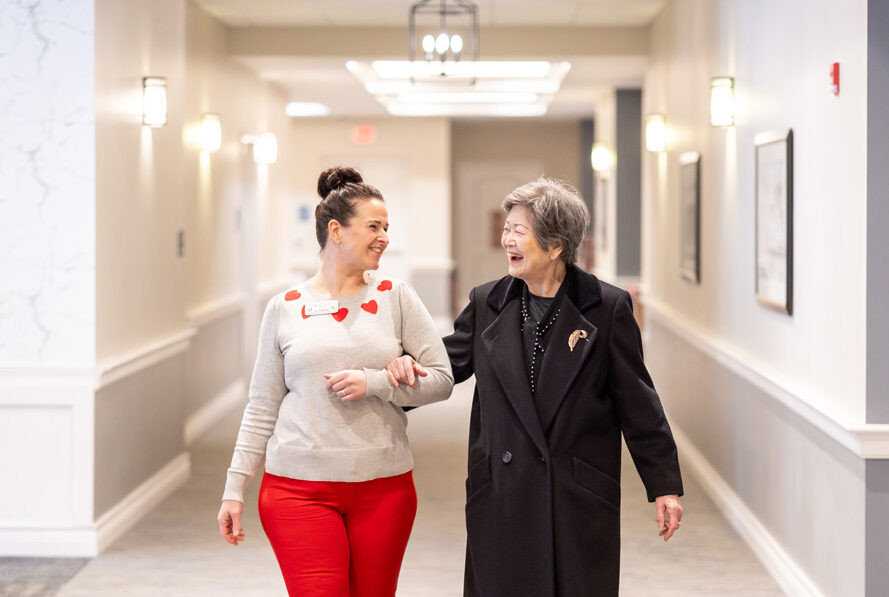 A senior resident enjoys a friendly walk with a team member through a brightly lit hallway. The team member wears a festive heart-themed sweater, sharing smiles and conversation.