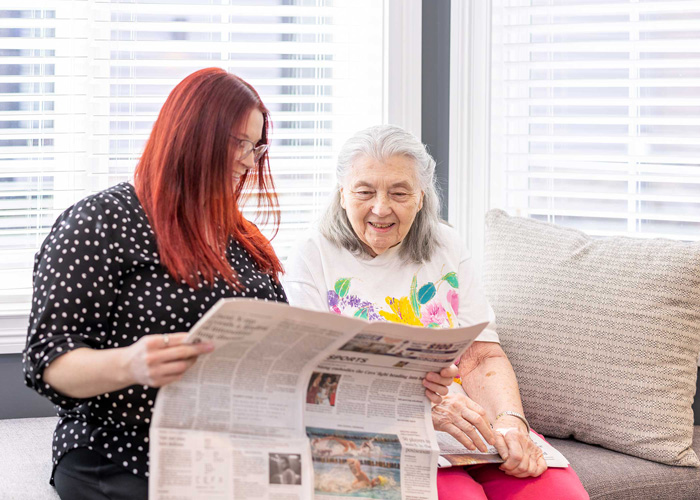 A young woman with red hair and glasses reads a newspaper with an senior resident in a floral shirt, both smiling and sitting together on a comfortable sofa by a window.