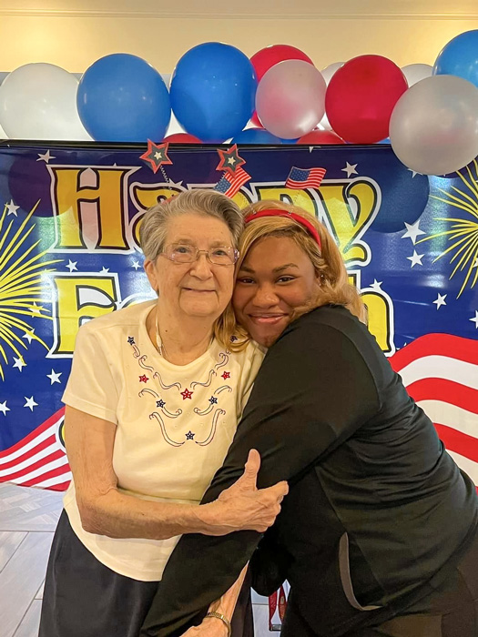 Senior resident and team member share a joyful embrace in front of a festive Fourth of July backdrop with red, white, and blue balloons, celebrating Independence Day together.