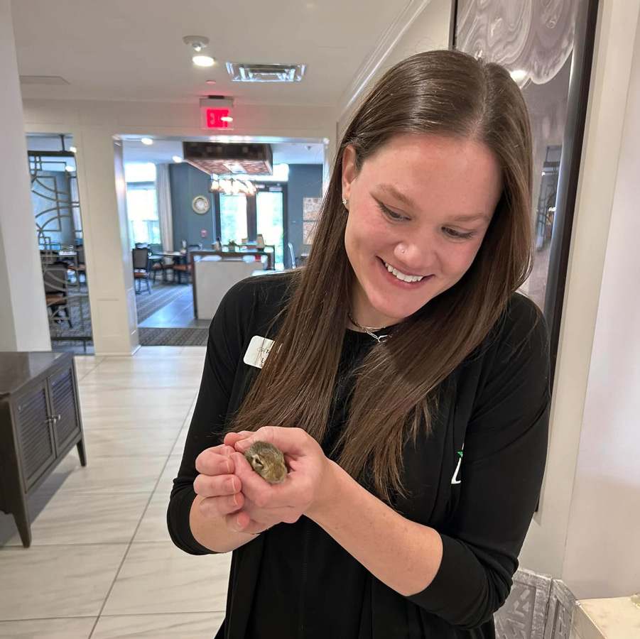 A smiling woman holding a small, furry animal in her hands, enjoying a visit from the furry friend inside a senior living community.