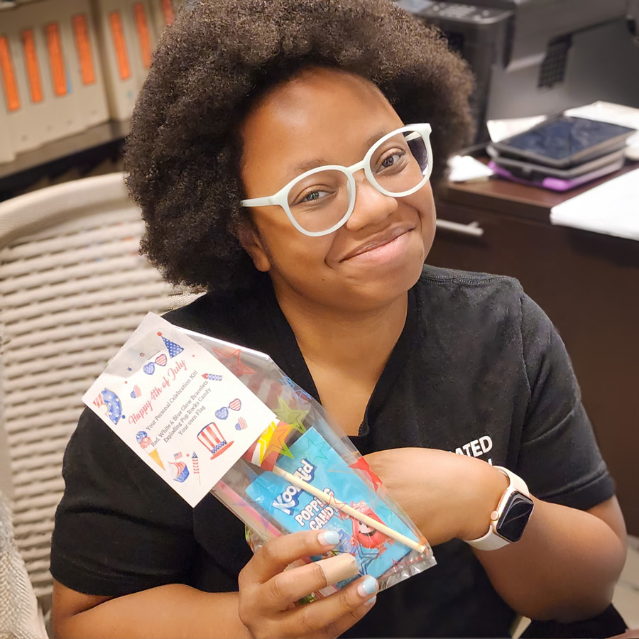 A woman with curly hair and glasses holding a festive 4th of July celebration pack, including Kool-Aid and Pop Rocks, while sitting at her desk.