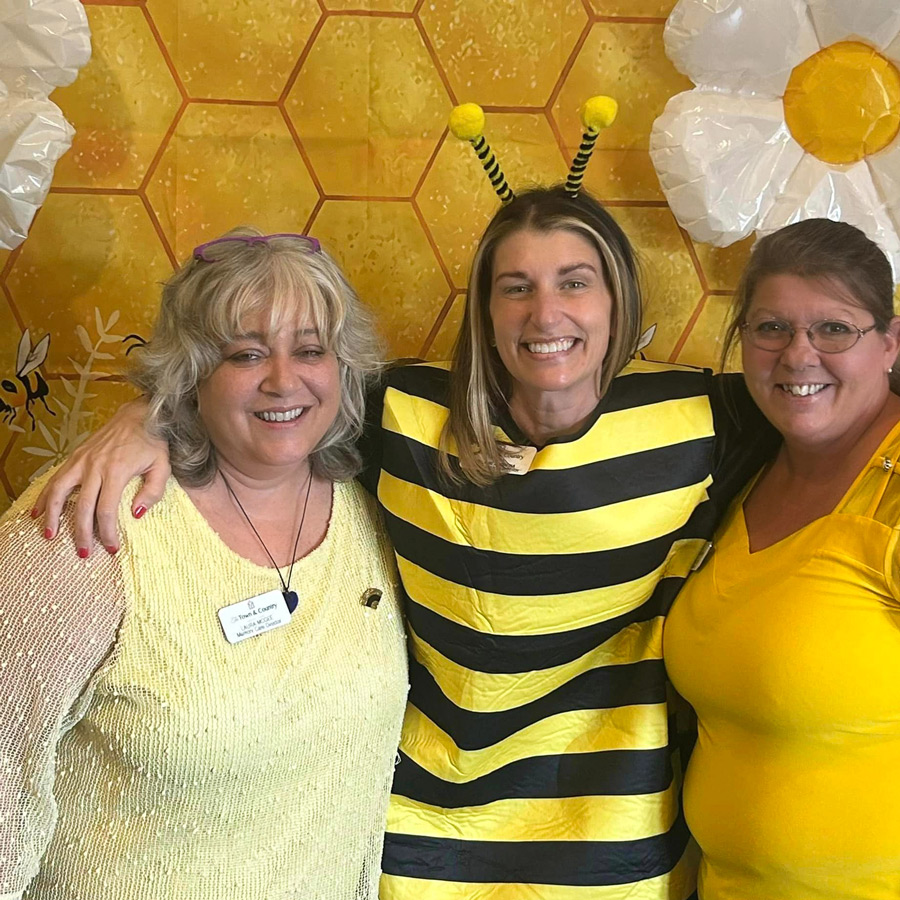 Three women smiling and dressed in bee-themed yellow and black outfits, including one in a bee costume with antennae, standing in front of a honeycomb-patterned backdrop during a senior living community event.