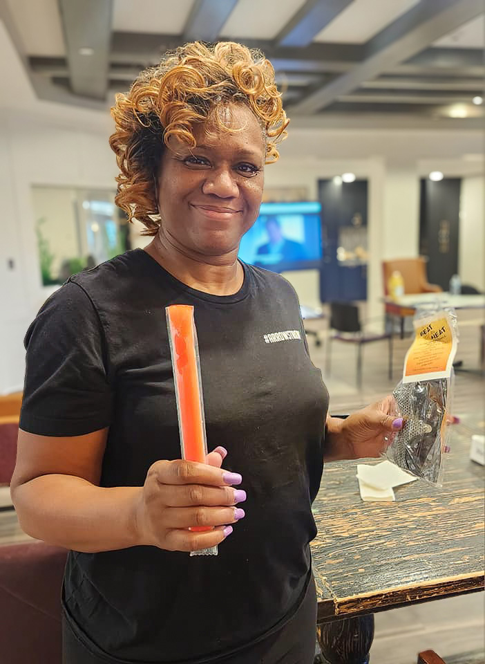 Employee in a black shirt with '#ARROWSTRONG' holding a popsicle and a gift bag labeled 'Beat the Heat' at a senior living community.