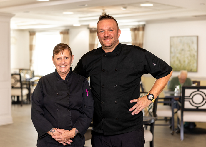 Two chefs in black uniforms stand smiling in a well-lit dining area, showcasing their culinary team. A senior resident can be seen seated at a table in the background.