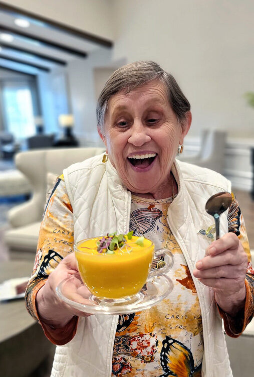 Smiling senior resident joyfully holds a bowl of vibrant orange soup garnished with greens, wearing a colorful butterfly-patterned shirt and white vest, ready to enjoy the meal.