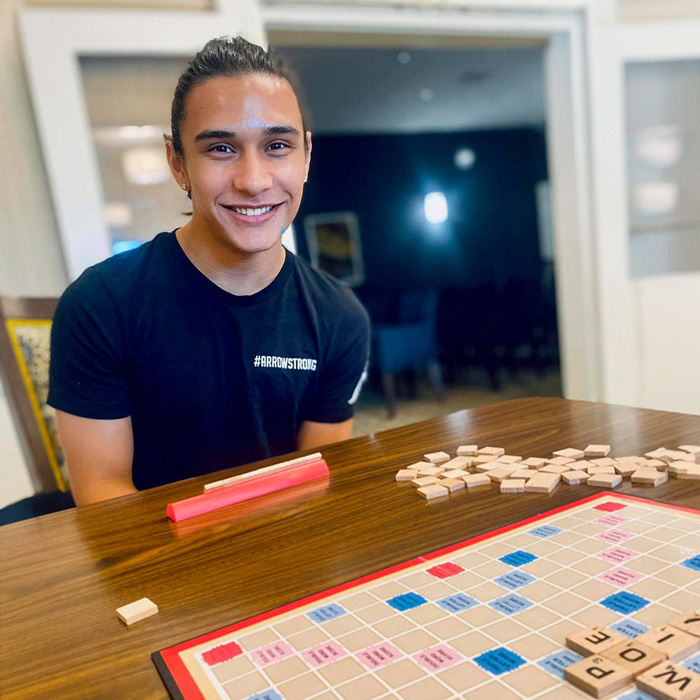 Smiling young man with a ponytail wearing a black '#ArrowStrong' shirt, sitting at a table with a Scrabble board game in progress and scattered tiles in a well-lit room.