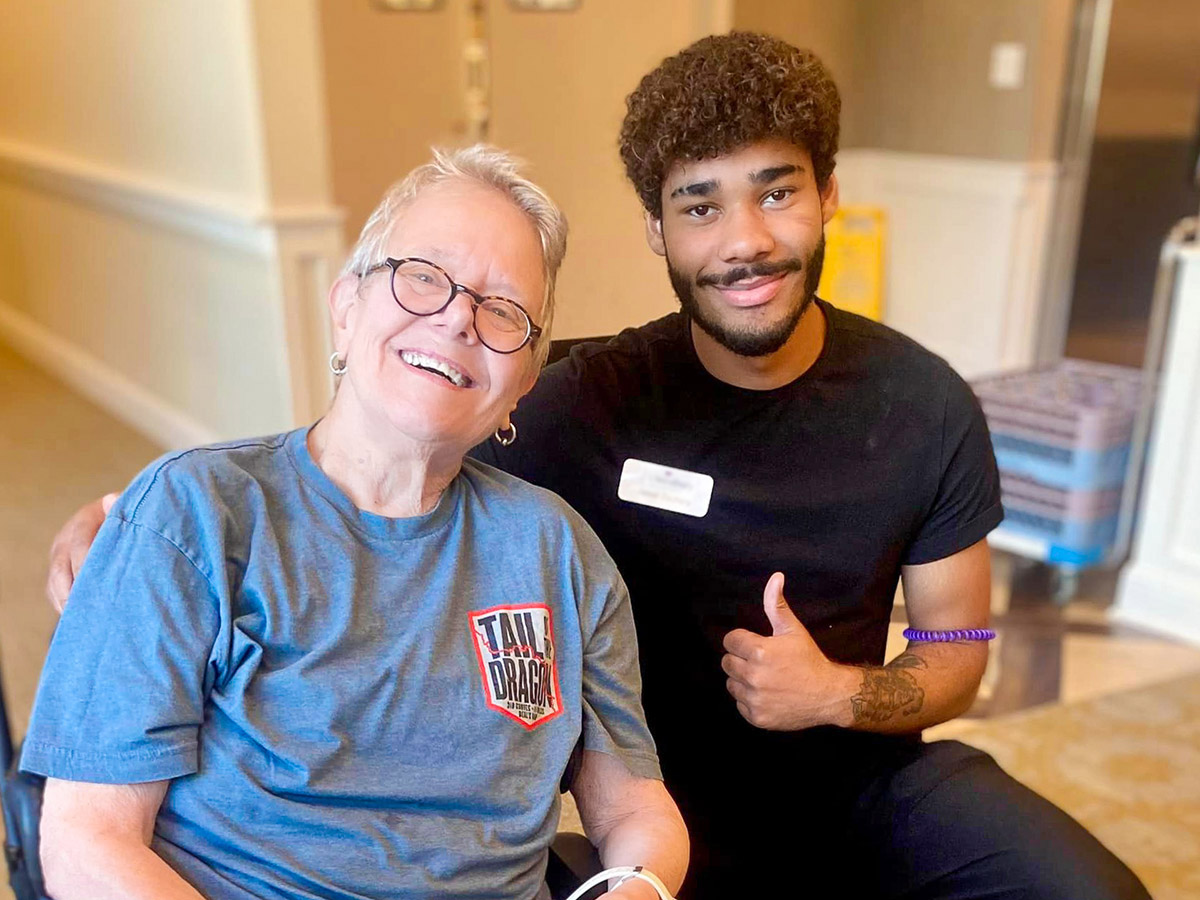 Smiling senior resident wearing glasses and a blue T-shirt sitting next to a young male staff member in a black shirt, both giving thumbs up, enjoying time together.