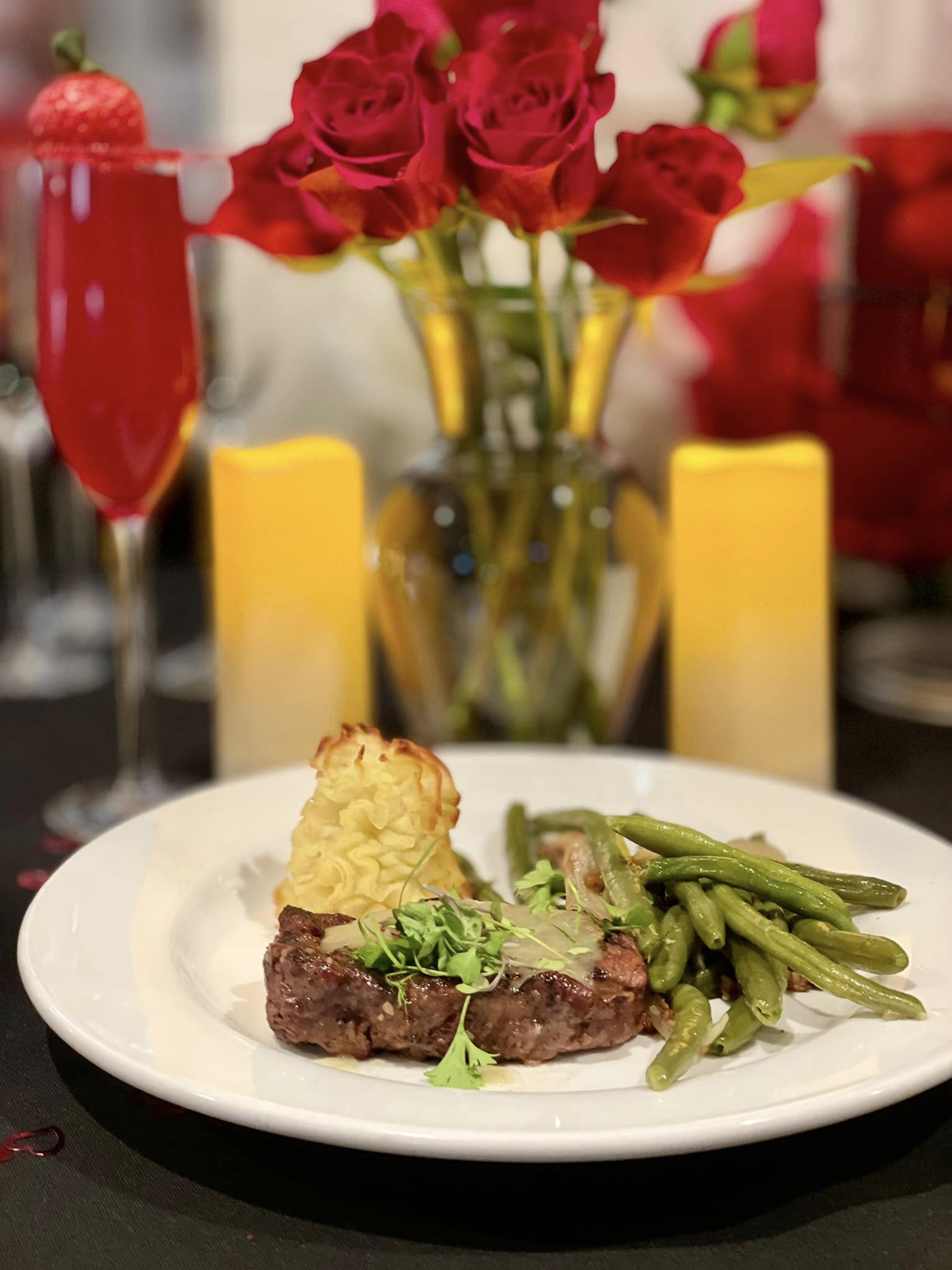 Elegant dinner featuring a perfectly cooked steak, green beans, and a duchess potato, accompanied by a festive drink and red roses in the background, set on a black tablecloth.