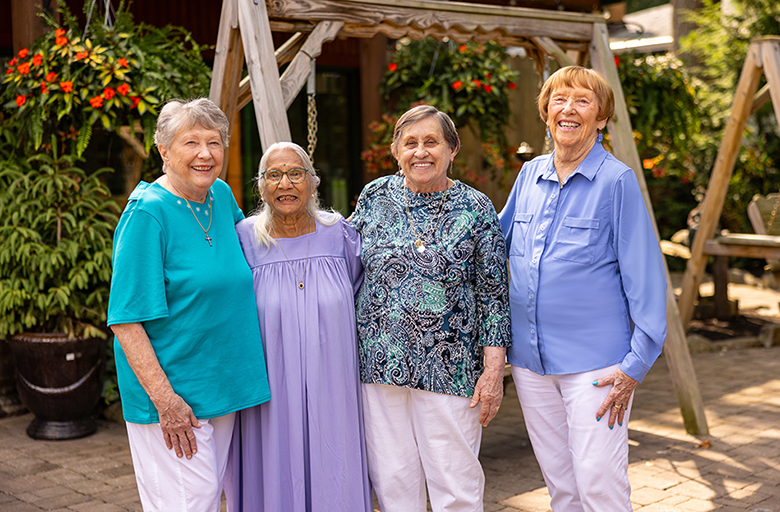 Four senior residents stand together outside, smiling warmly. The garden setting features a swing and vibrant flowers in the background, creating a serene and welcoming atmosphere.