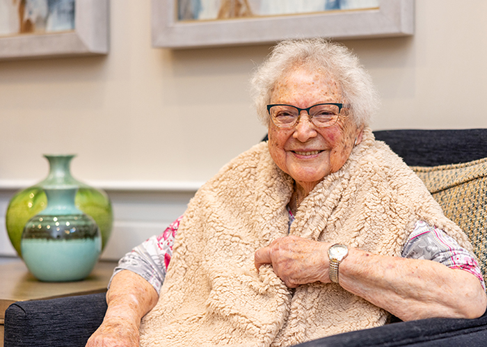 Senior resident sitting comfortably in a chair, wrapped in a cozy beige vest. She is smiling warmly, wearing glasses, and a watch on her wrist, with vases in the background.