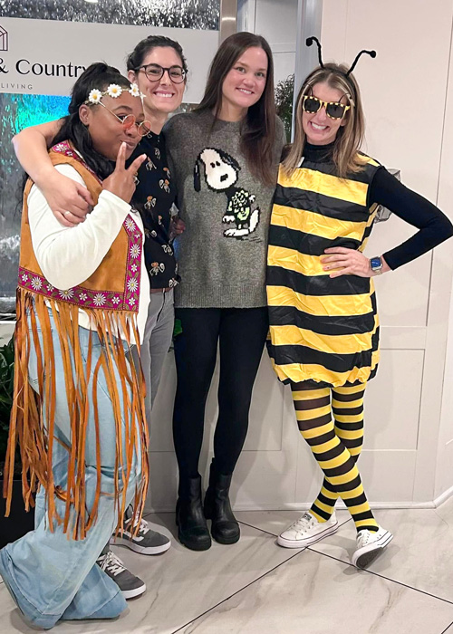 A cheerful group of four women dressed in festive costumes, including a flower child, a Snoopy sweater, and a bee outfit, posing with smiles and laughter in a senior living community, fostering a sense of joy and camaraderie.