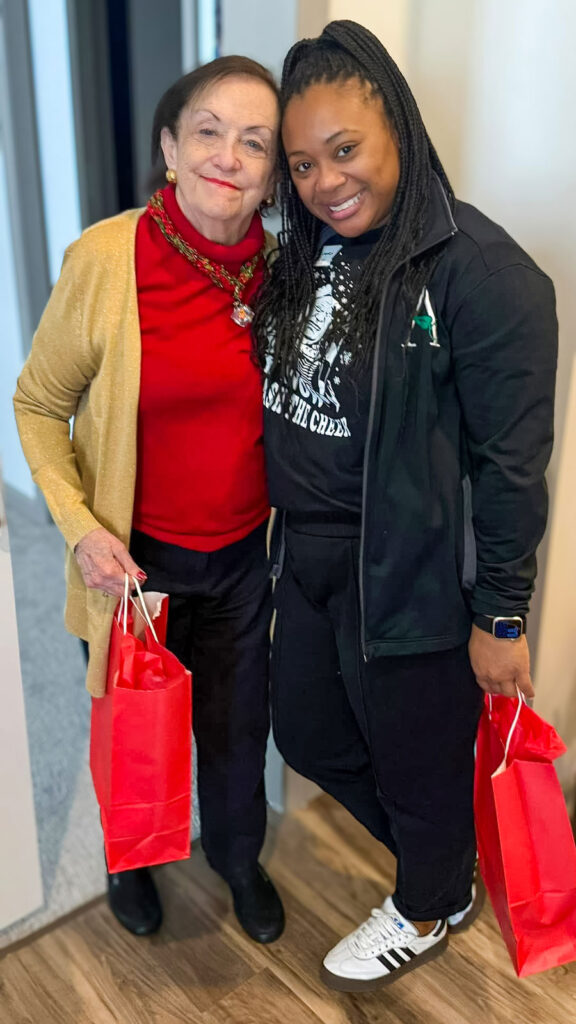 A vibrant image featuring a senior resident in a red sweater and gold cardigan standing arm-in-arm with a staff member dressed in black, both holding bright red gift bags, radiating warmth and holiday cheer.